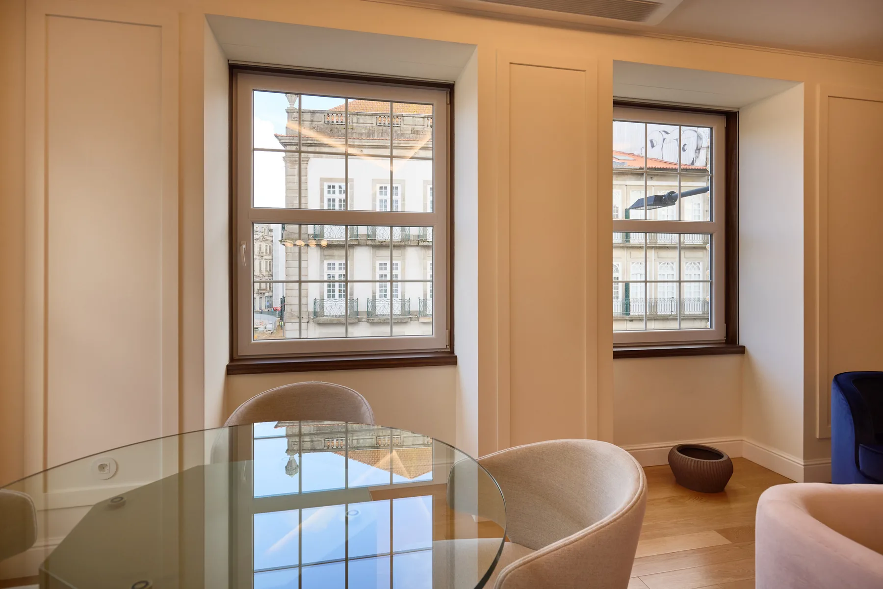 Dining area with a glass table and two windows overlooking nearby buildings.
