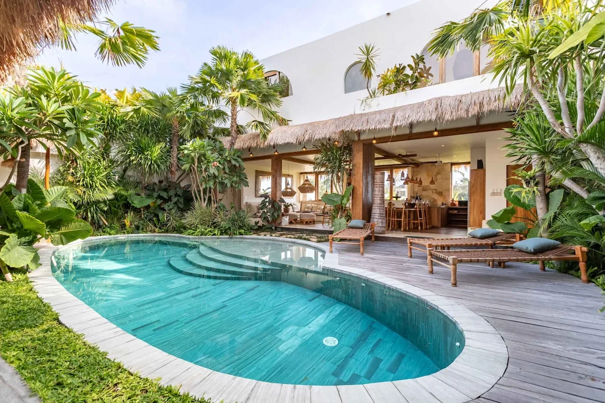 A swimming pool sits beside a shaded patio with seating and tropical plants.