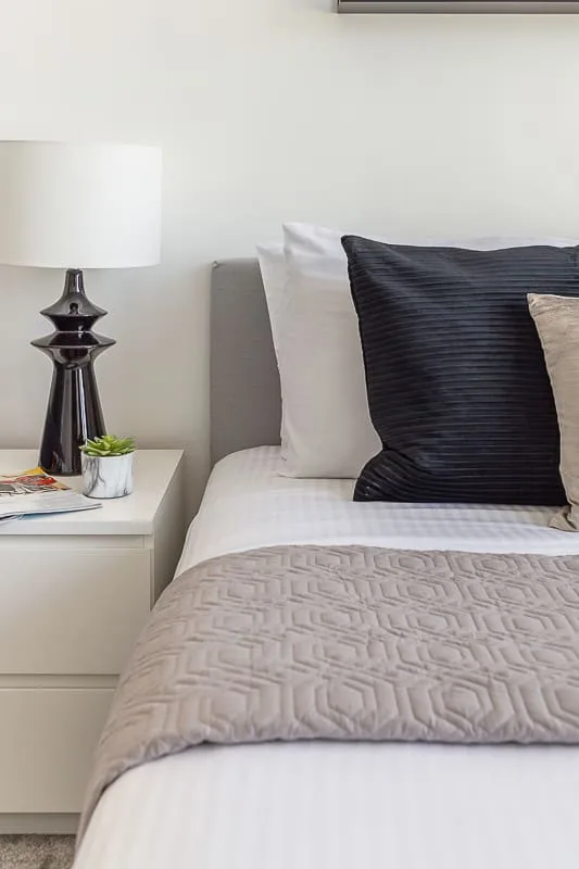 A neatly made bed with textured bedding sits beside a white bedside table and lamp.