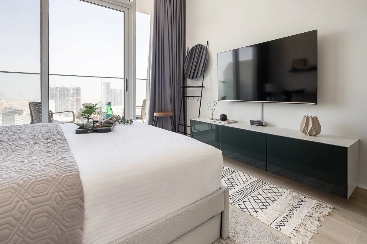 A bright bedroom with a large bed beside floor-to-ceiling windows and a wall-mounted television.