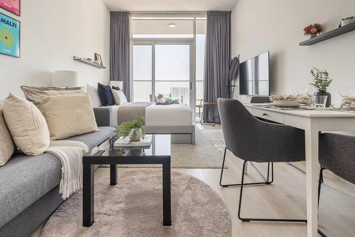 A bright studio room combines a seating area, bed, and desk by a large window.