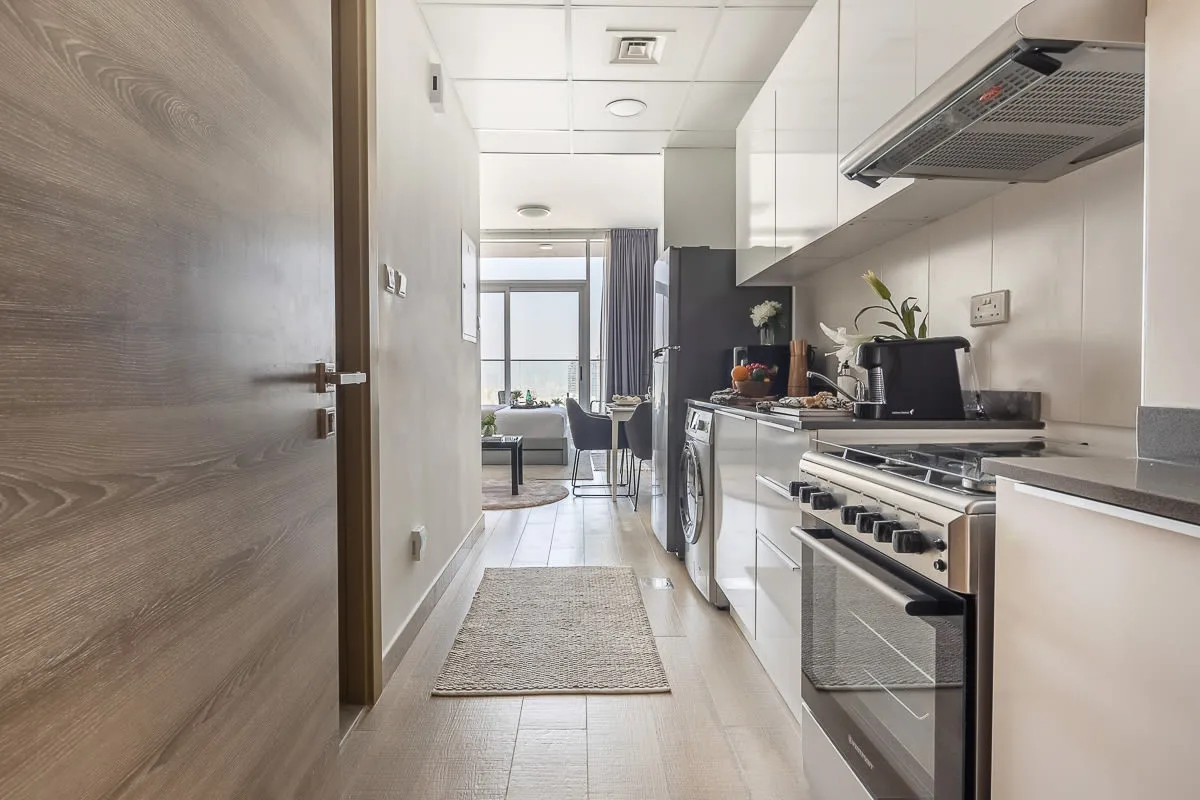 A narrow kitchen with white cabinets, a stove, and a dining area by the windows.