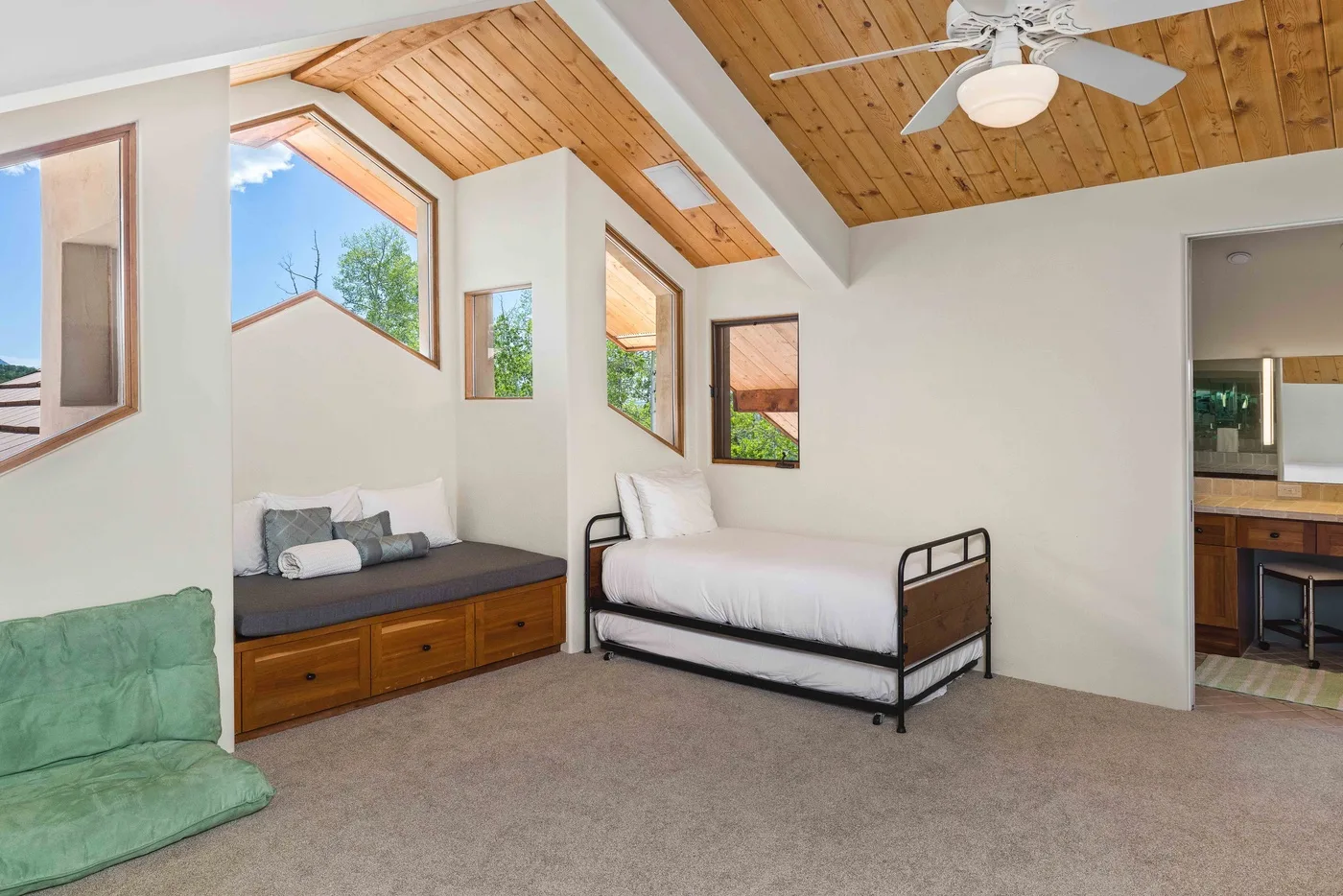 Un dormitorio luminoso con una cama, un asiento junto a la ventana y techo de madera inclinado.