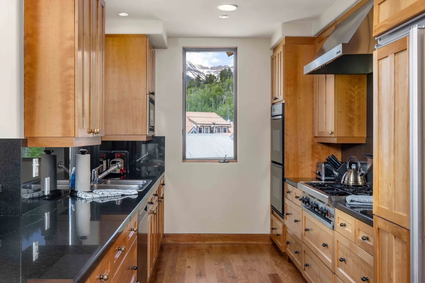 Una cocina en forma de pasillo con gabinetes de madera, encimeras oscuras y una ventana al fondo.