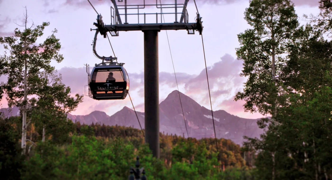 Un teleférico pasa sobre un paisaje arbolado con montañas al fondo.