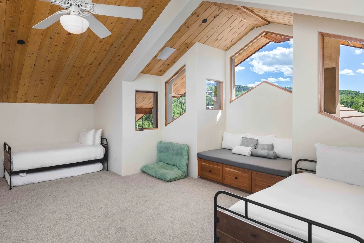 Un dormitorio abuhardillado y luminoso con varias camas, un asiento verde y ventanas amplias.