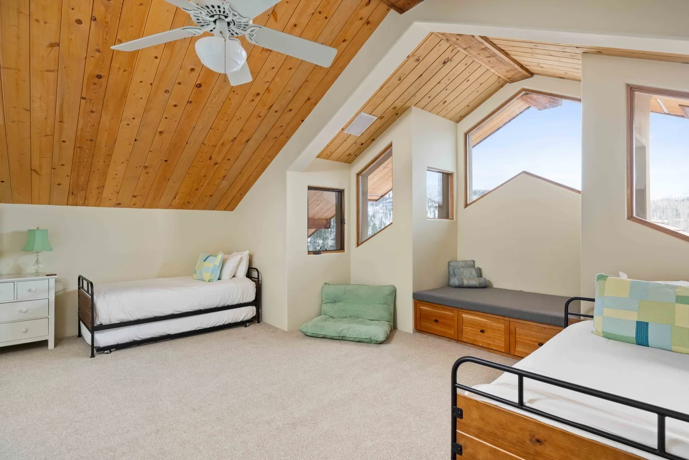 Un dormitorio luminoso en el ático, con techo inclinado de madera, camas y ventanas grandes.