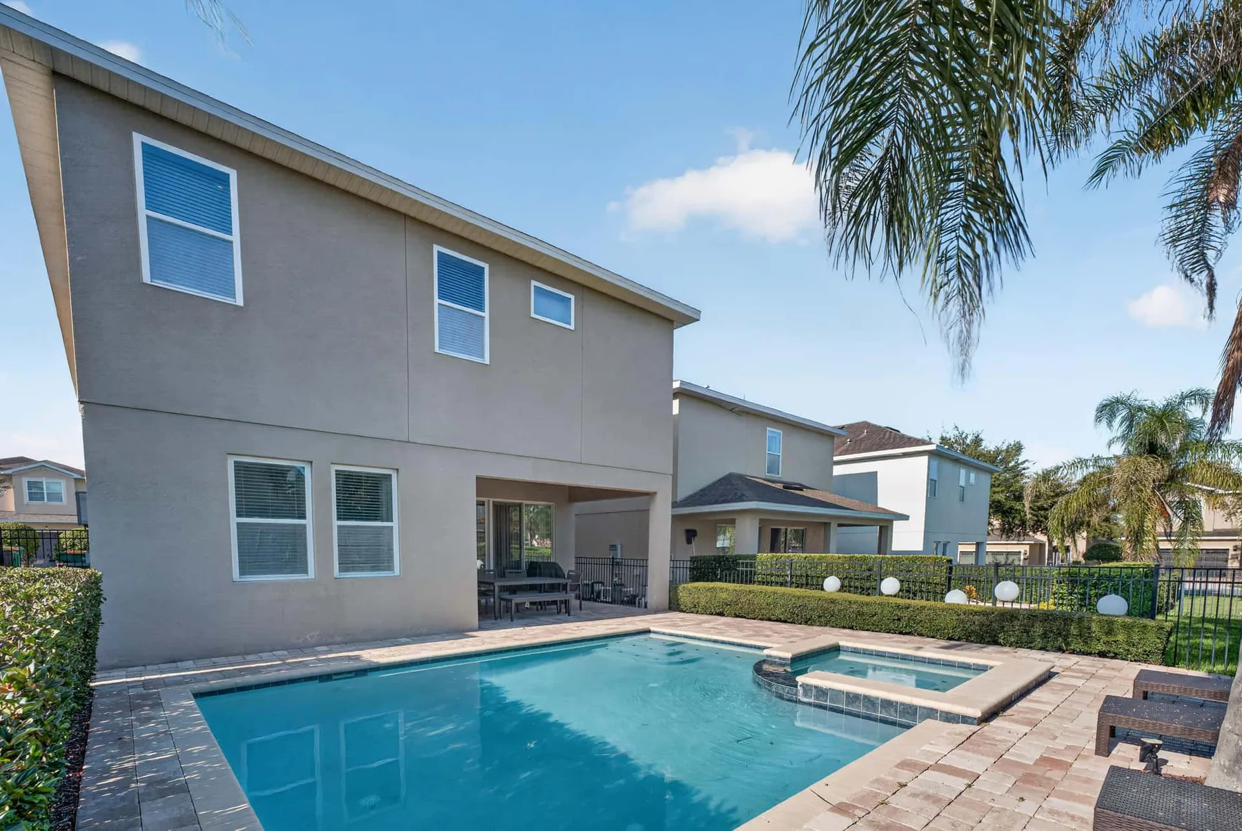 A backyard pool beside a two-story house, with patio seating and palm trees.