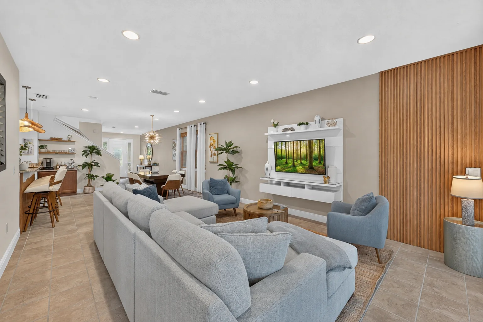 An open-plan living room with seating, a wall-mounted television, and a kitchen beyond.