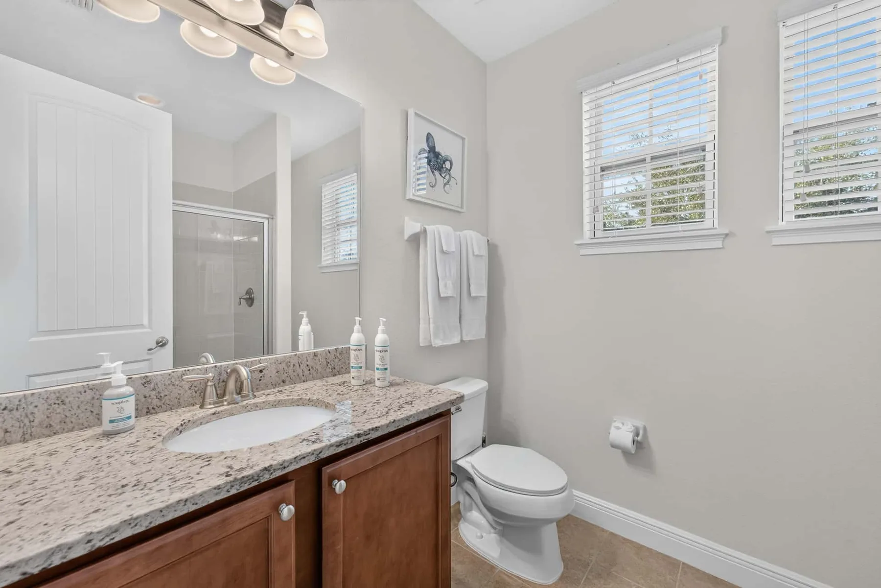 A bright bathroom with a vanity, toilet, and windows with blinds.