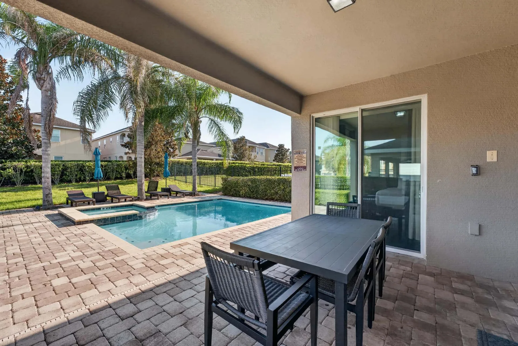 A covered patio with a dining table overlooks a swimming pool and palm trees.