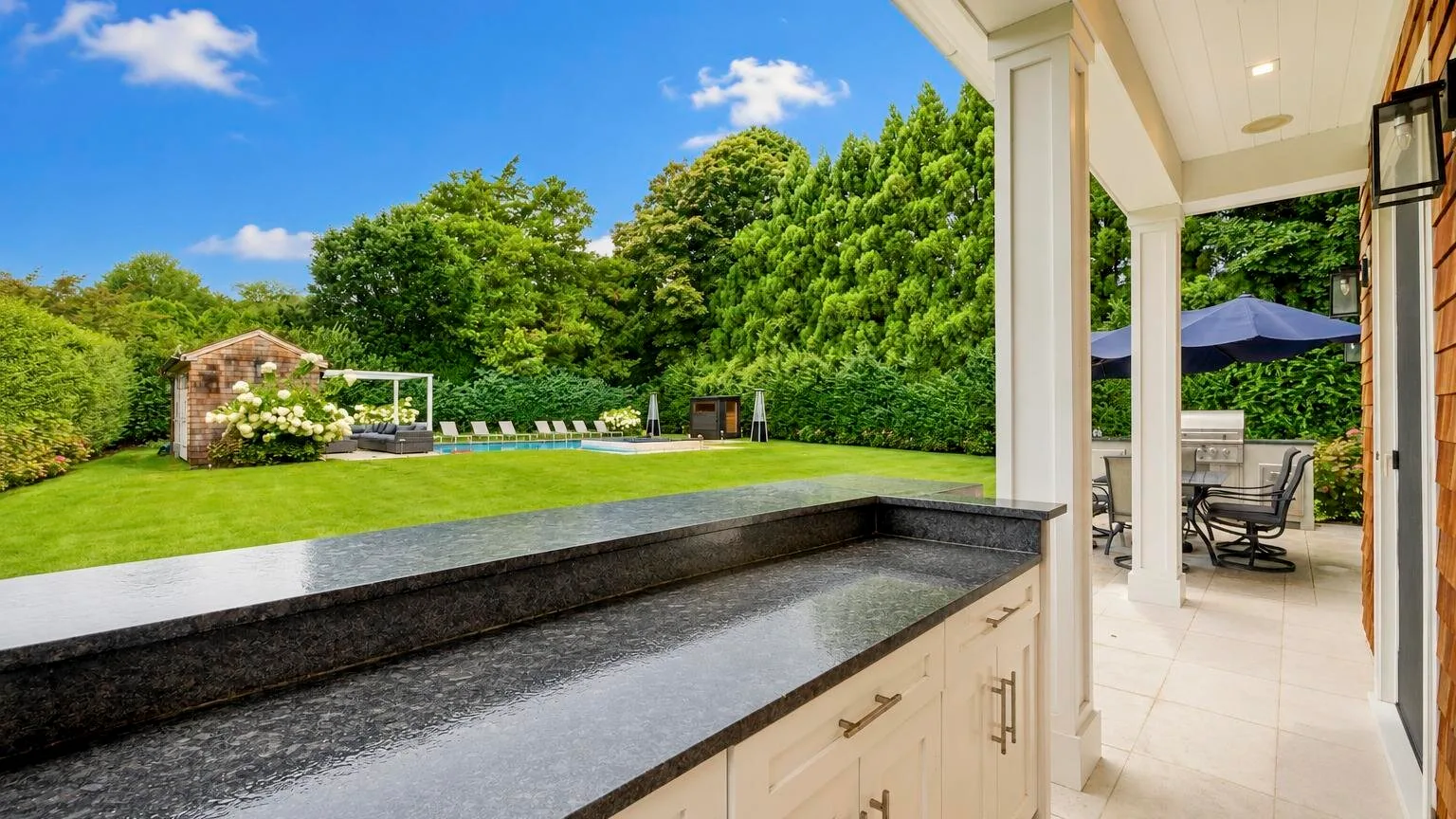 An outdoor kitchen counter overlooks a green lawn and a covered patio with seating.