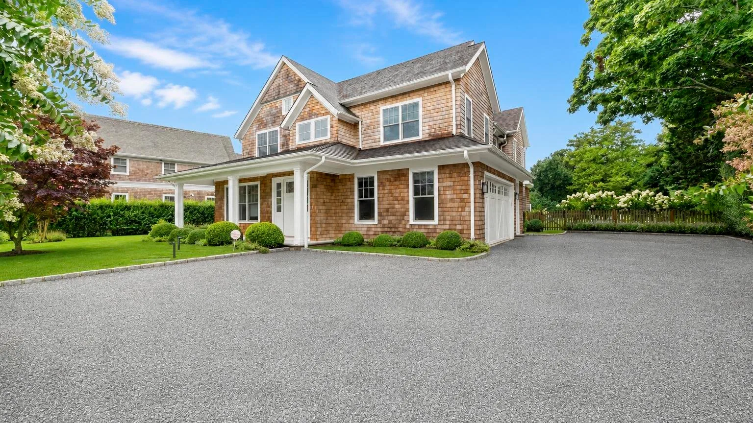 A shingled house stands beside a broad paved driveway, with trees and lawn.