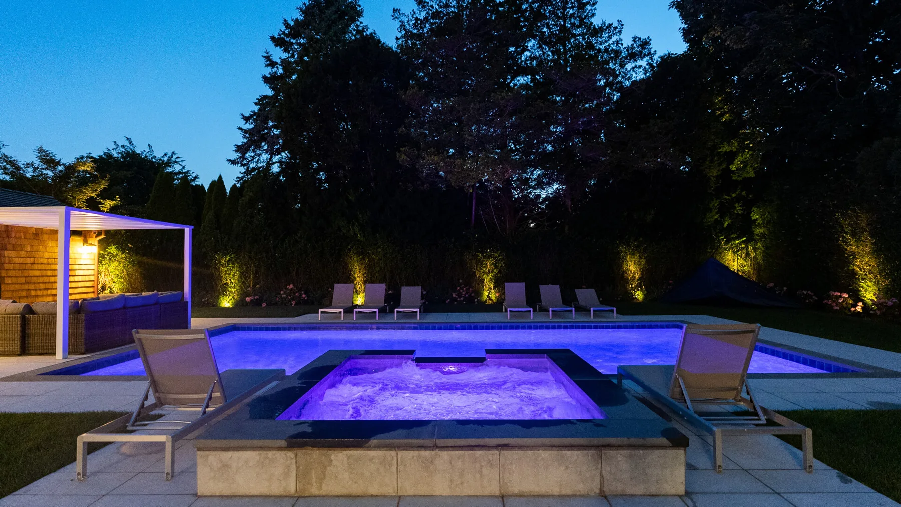 An outdoor pool and illuminated spa sit beside lounge seating at dusk.