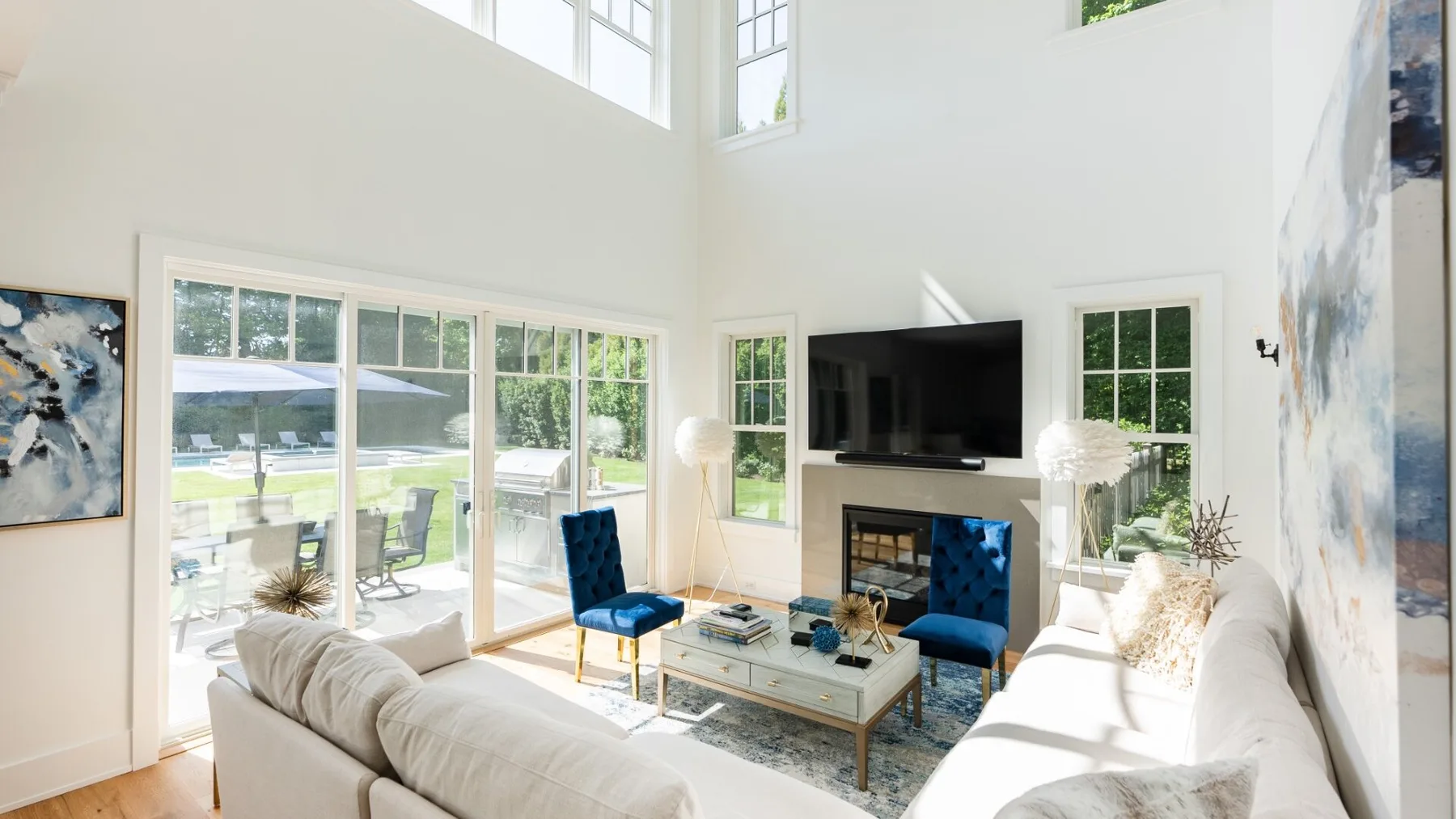 A bright living room with seating, a television, and large windows overlooking greenery.