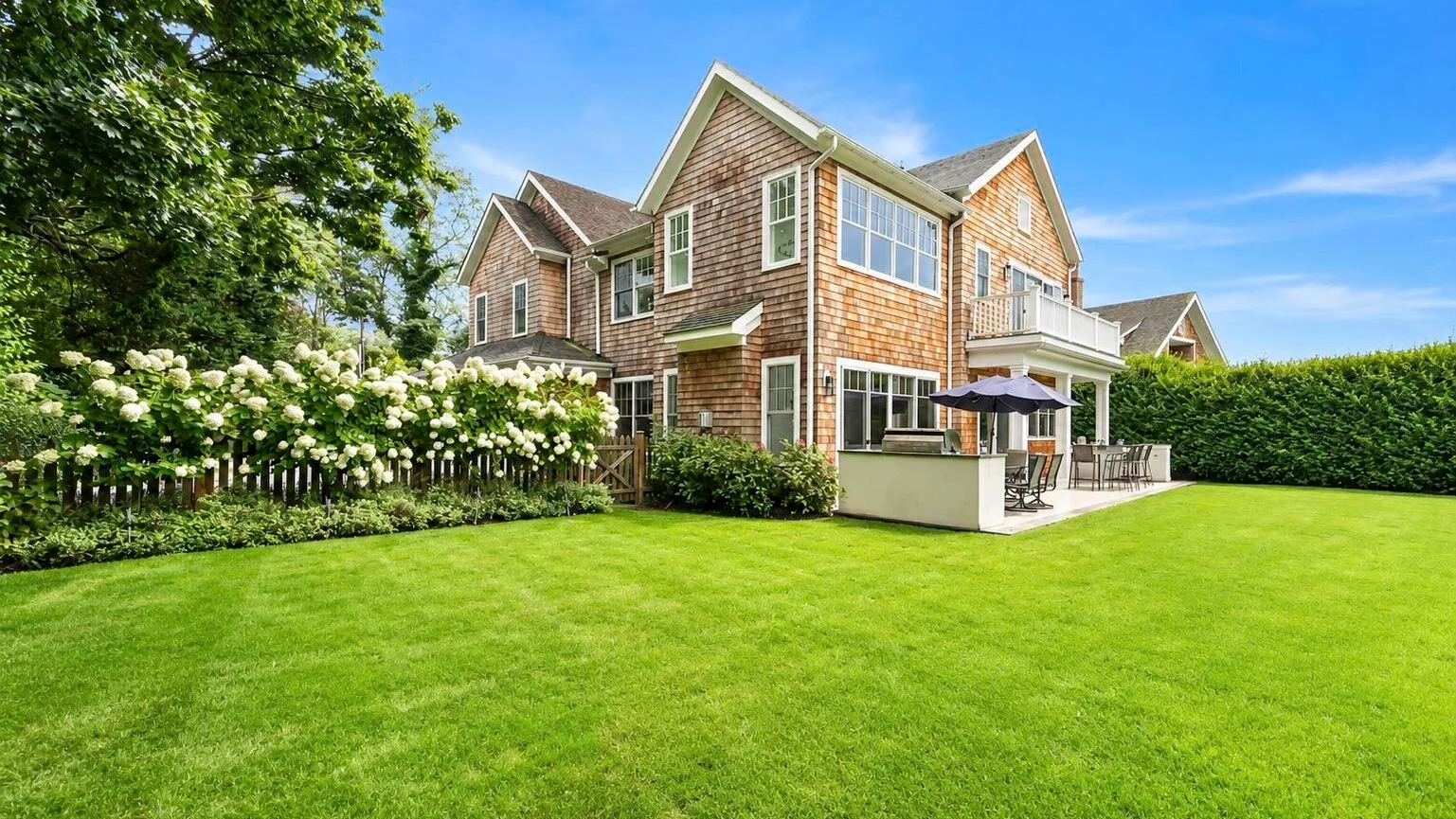 A large house stands beside a broad green lawn and flowering shrubs.