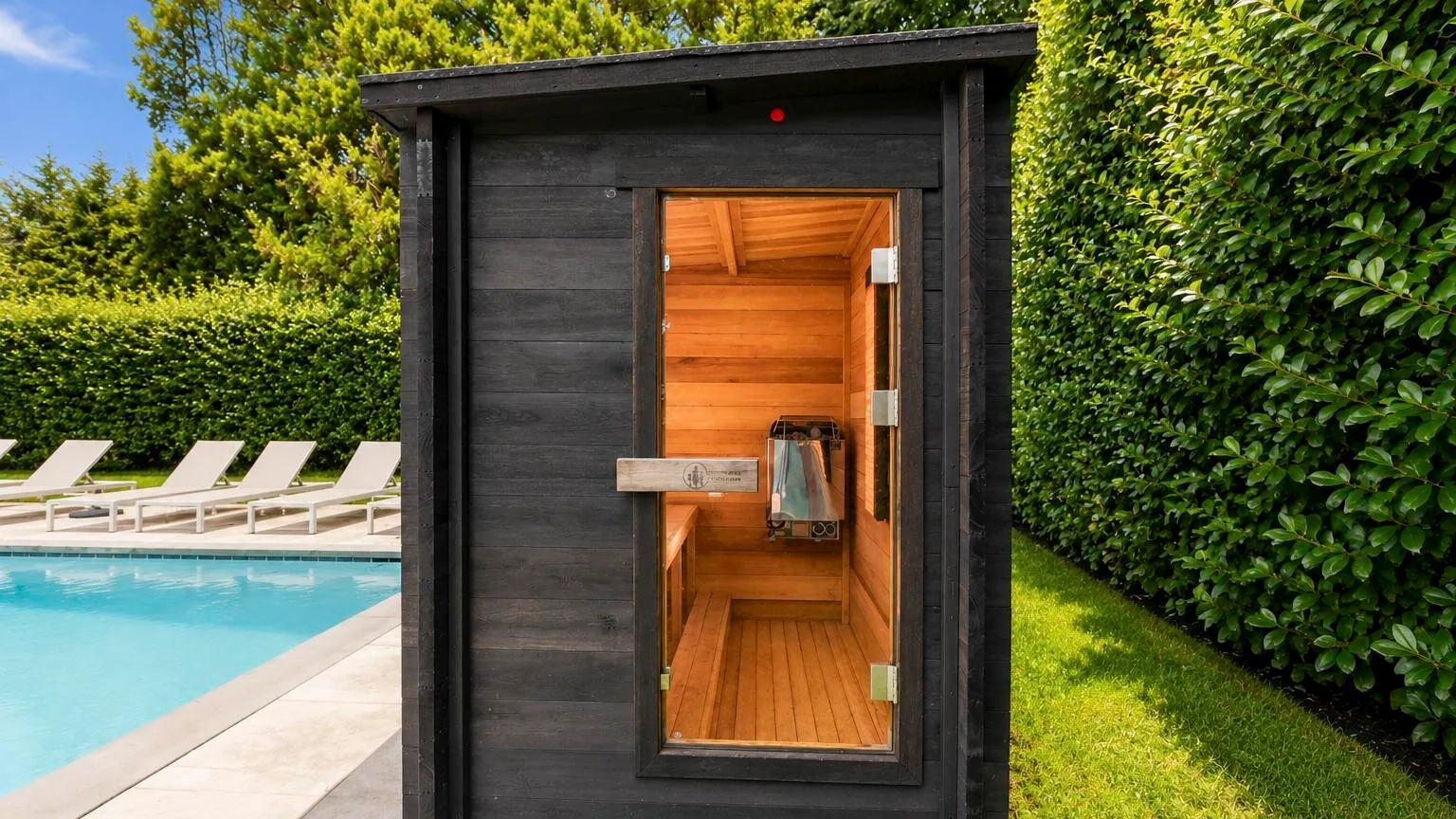 A wooden sauna stands beside an outdoor swimming pool and green hedges.