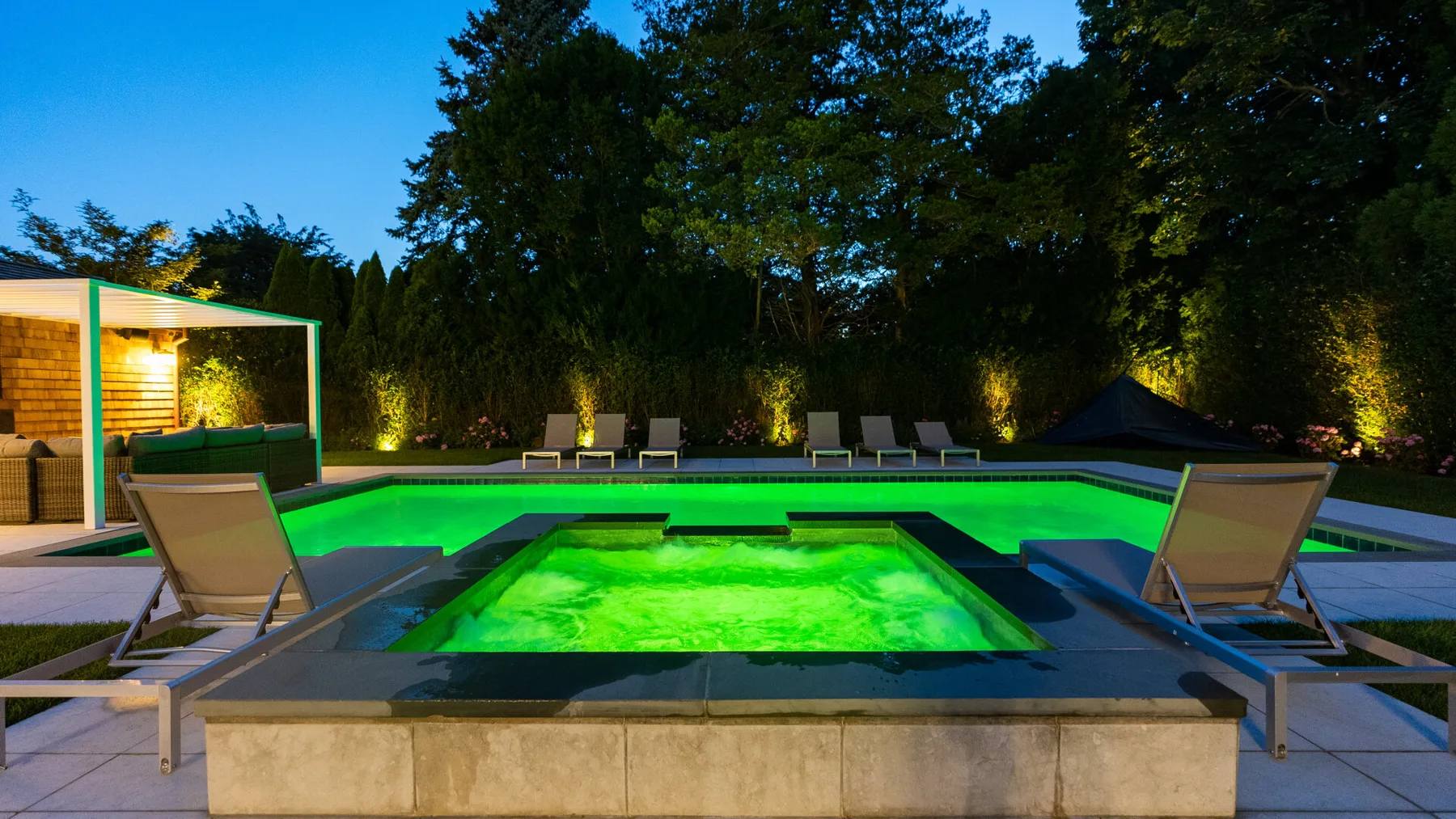 An illuminated outdoor pool and spa area at dusk, surrounded by trees.