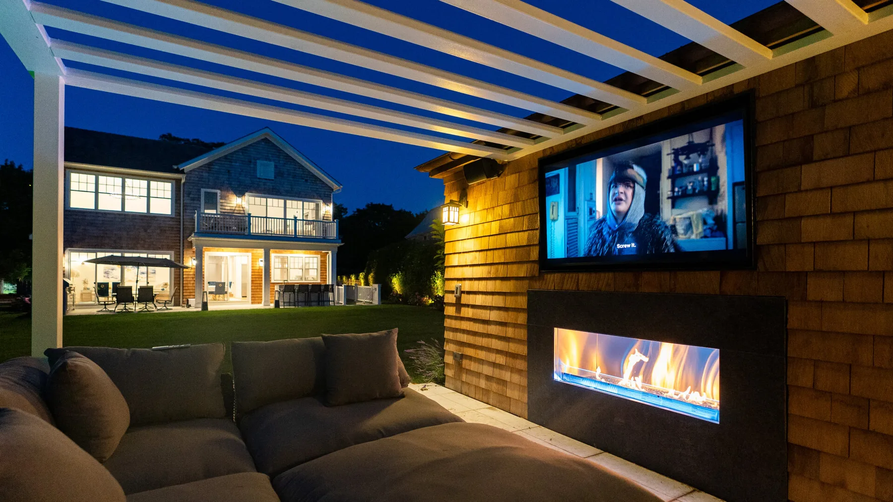 An outdoor lounge with seating and a wall-mounted television beside a lit fireplace.