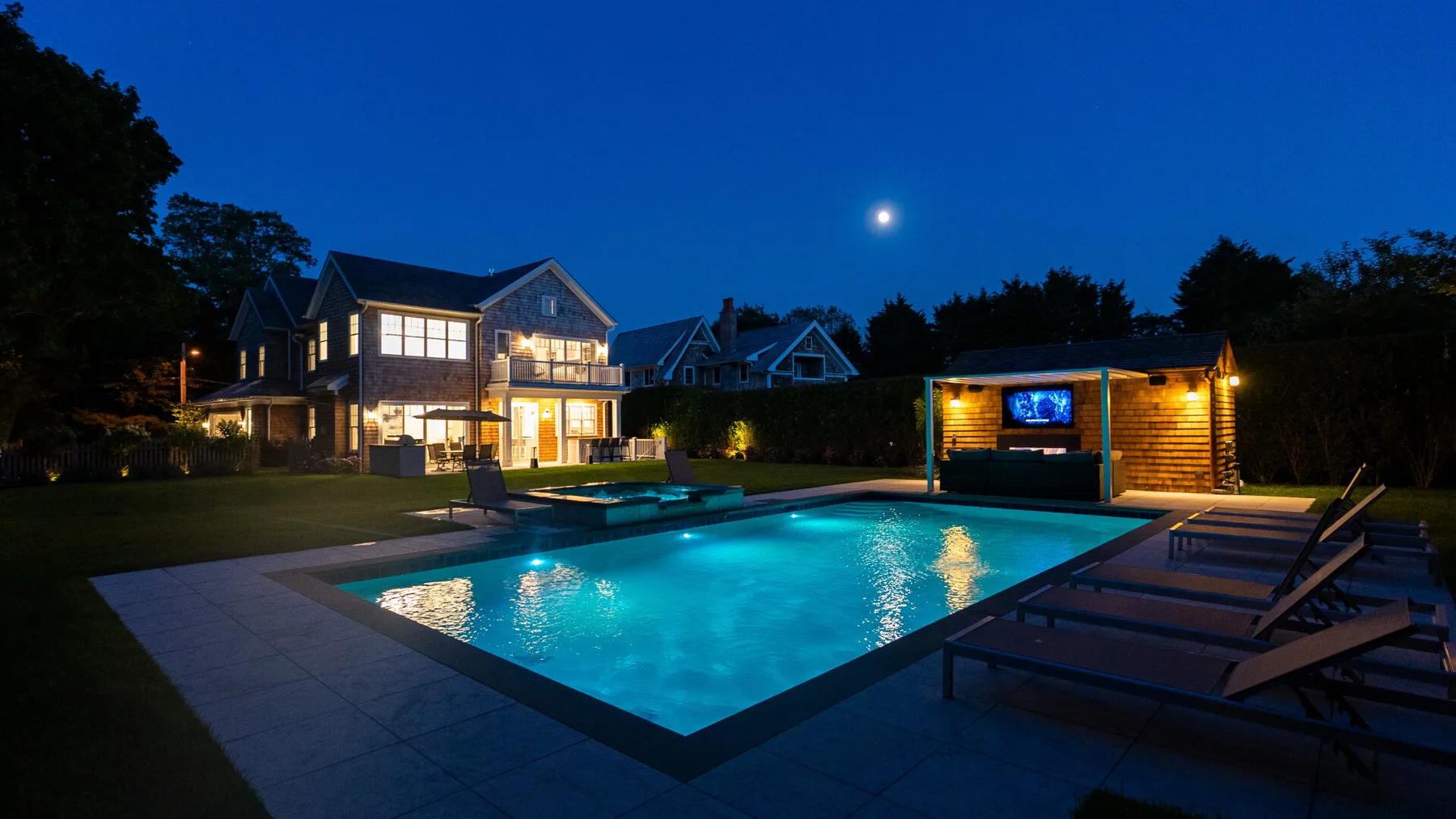 A lit swimming pool sits beside an illuminated house at dusk.