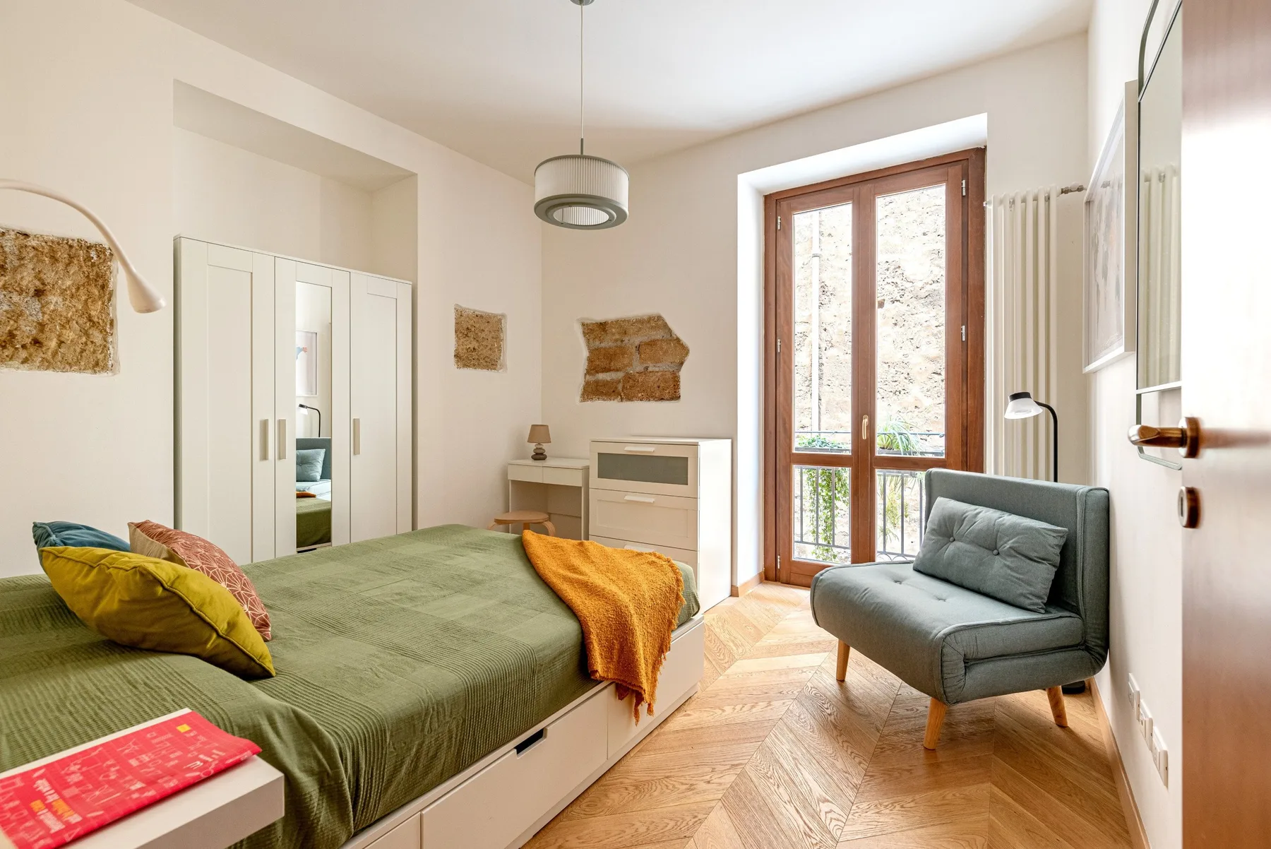 A bedroom with a green bedspread, a gray seat, and tall windows.