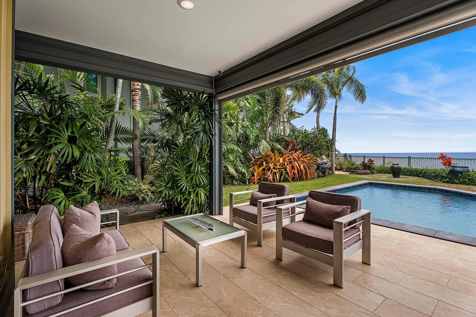 A covered patio seating area overlooks a swimming pool and lush tropical landscaping.