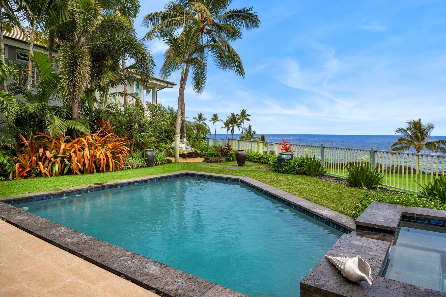 A swimming pool beside a garden, with palm trees and the ocean beyond.