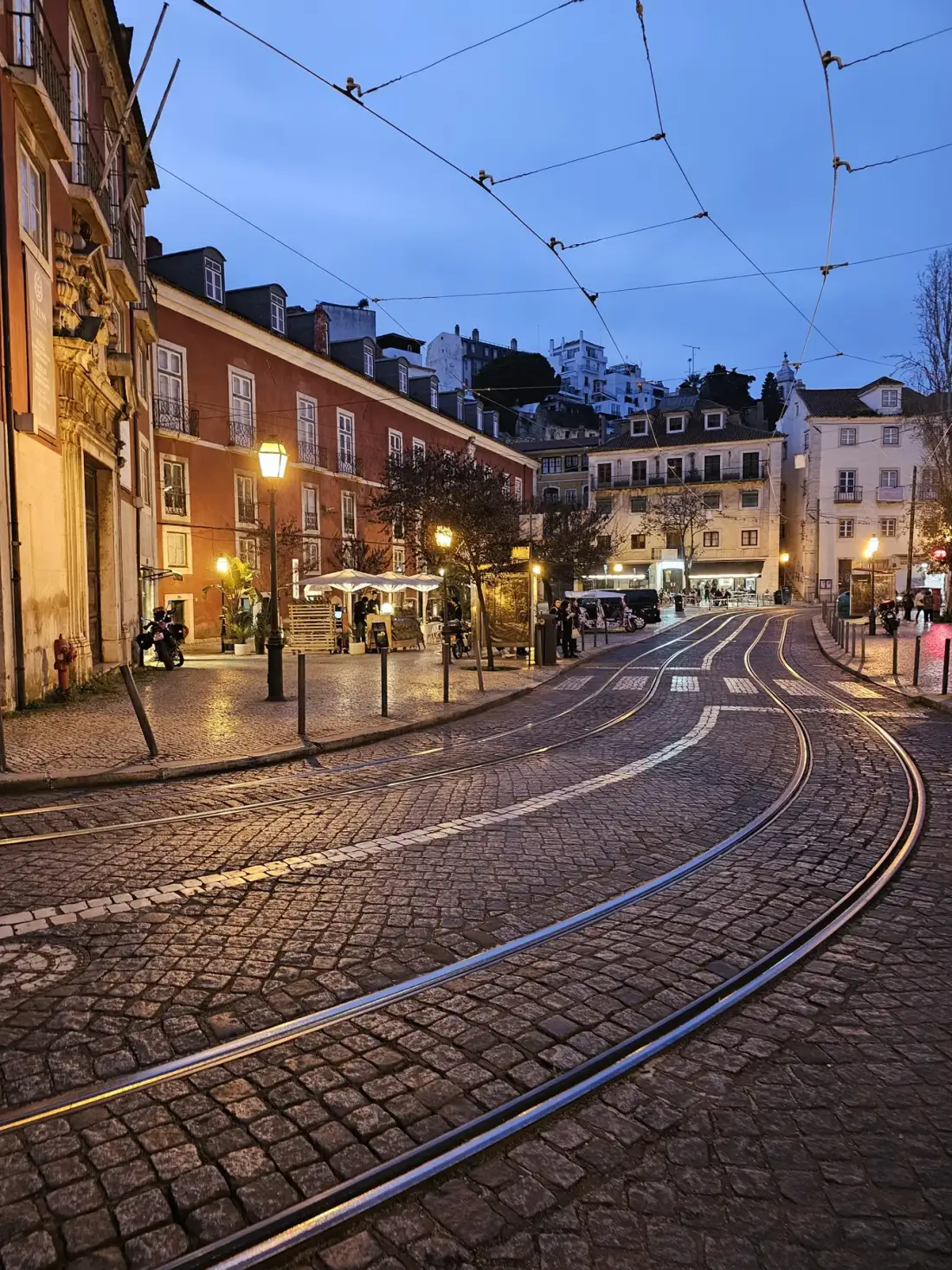 Una calle empedrada se curva entre edificios históricos bajo cables de tranvía al anochecer.
