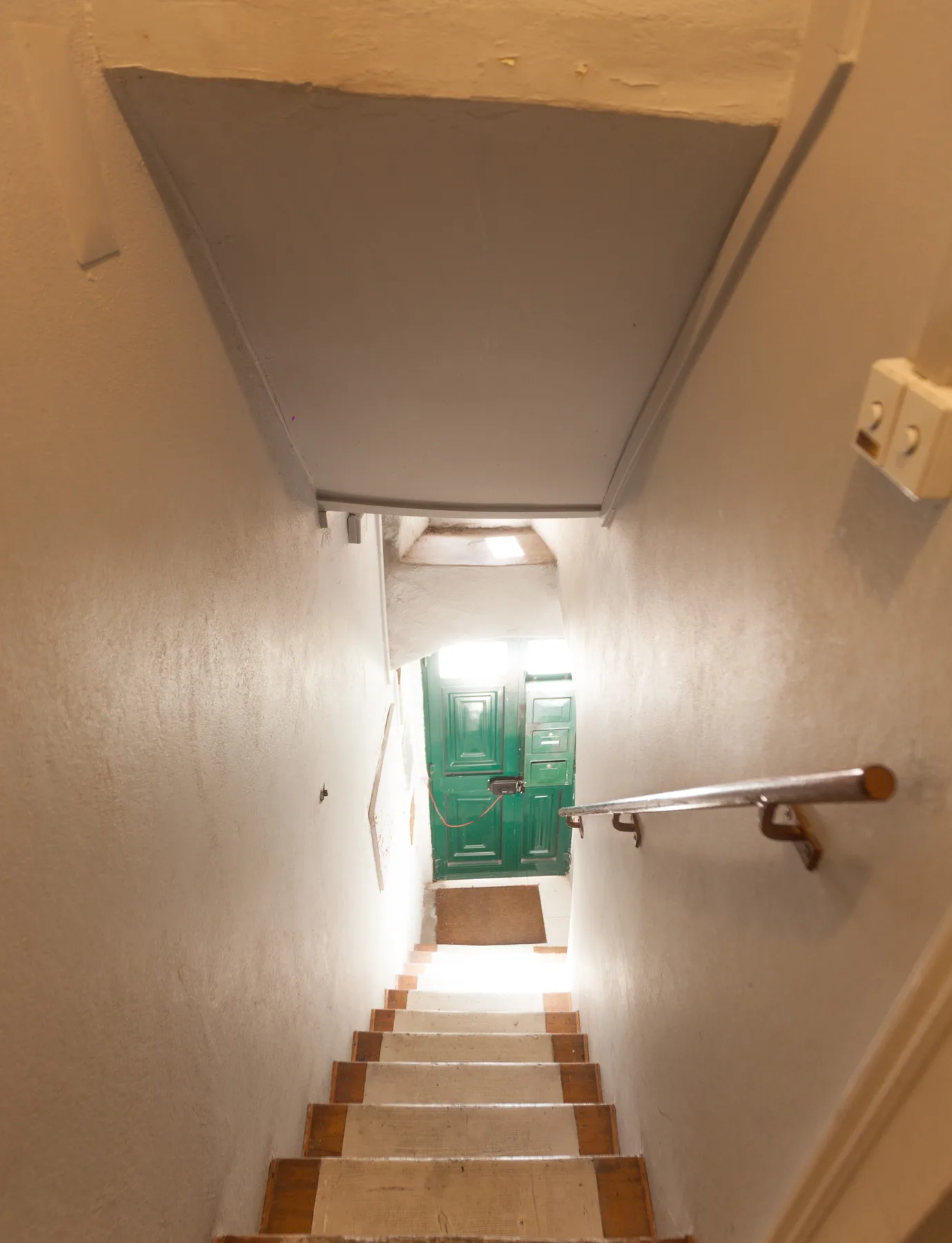 Una escalera estrecha baja hacia una puerta verde, con un pasamanos junto a la pared.