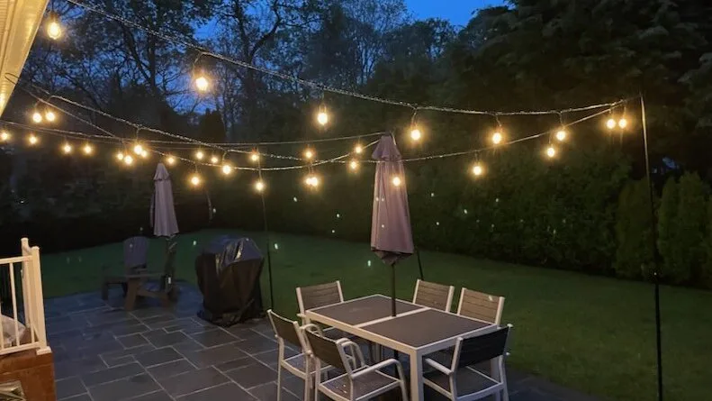 Un patio al aire libre con una mesa de comedor bajo luces colgantes, junto al césped.