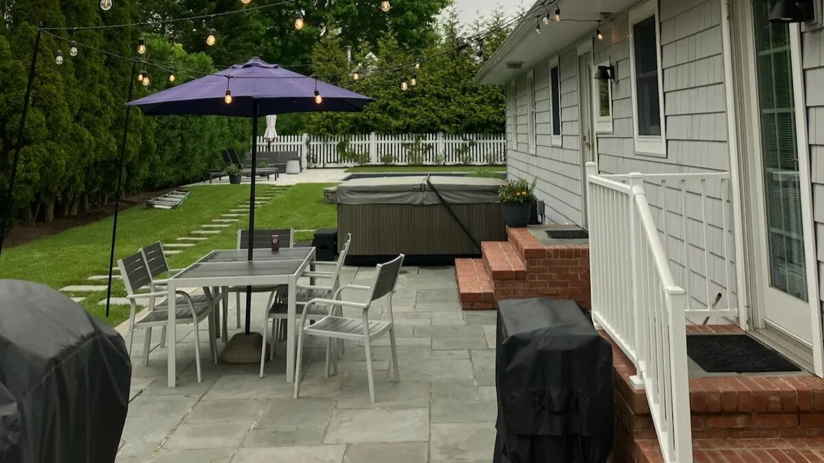 Un patio exterior pavimentado tiene asientos bajo una sombrilla junto a una casa.