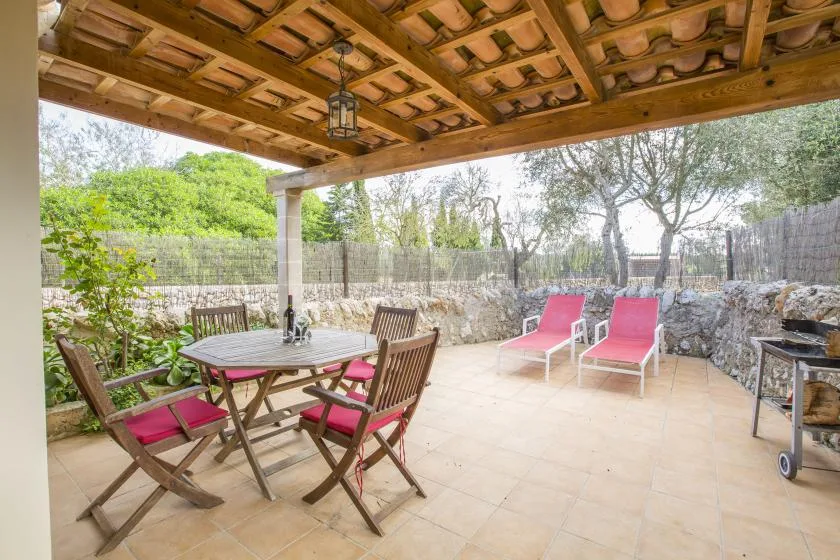 A covered patio with a dining table, outdoor seating, and trees beyond a stone wall.