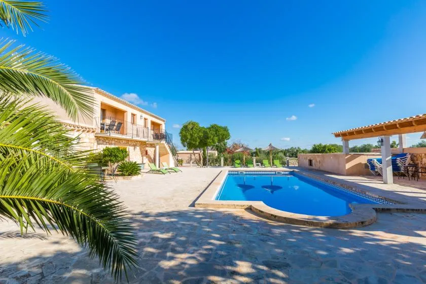 A swimming pool beside a sunlit villa and covered outdoor seating.