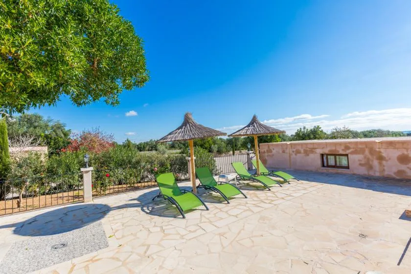 A sunny stone terrace has green loungers beneath two thatched parasols.