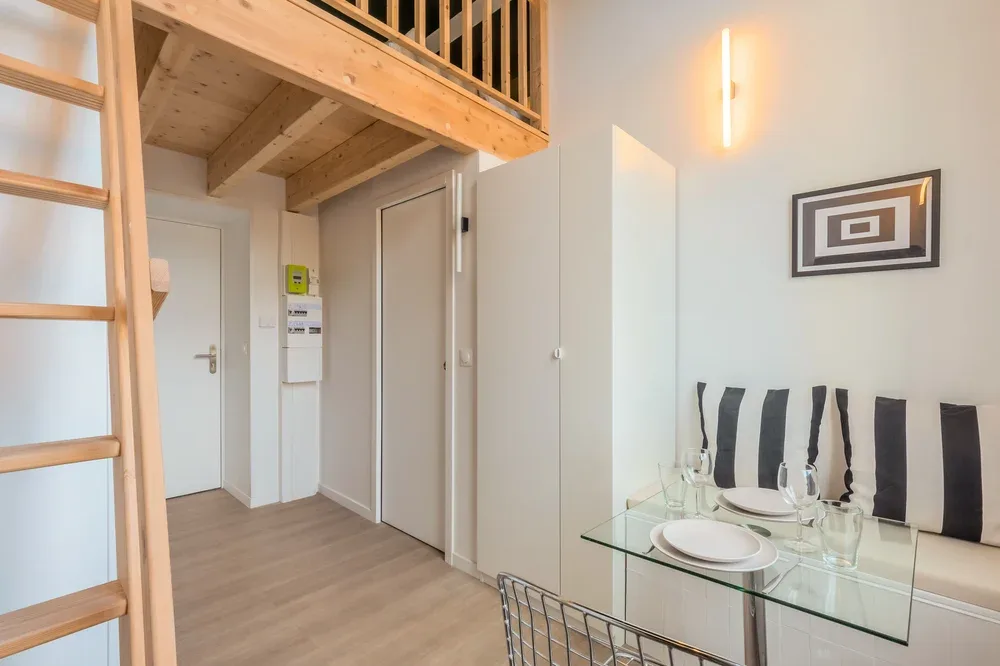 A dining area with a glass table and striped chairs sits beneath a wooden loft.