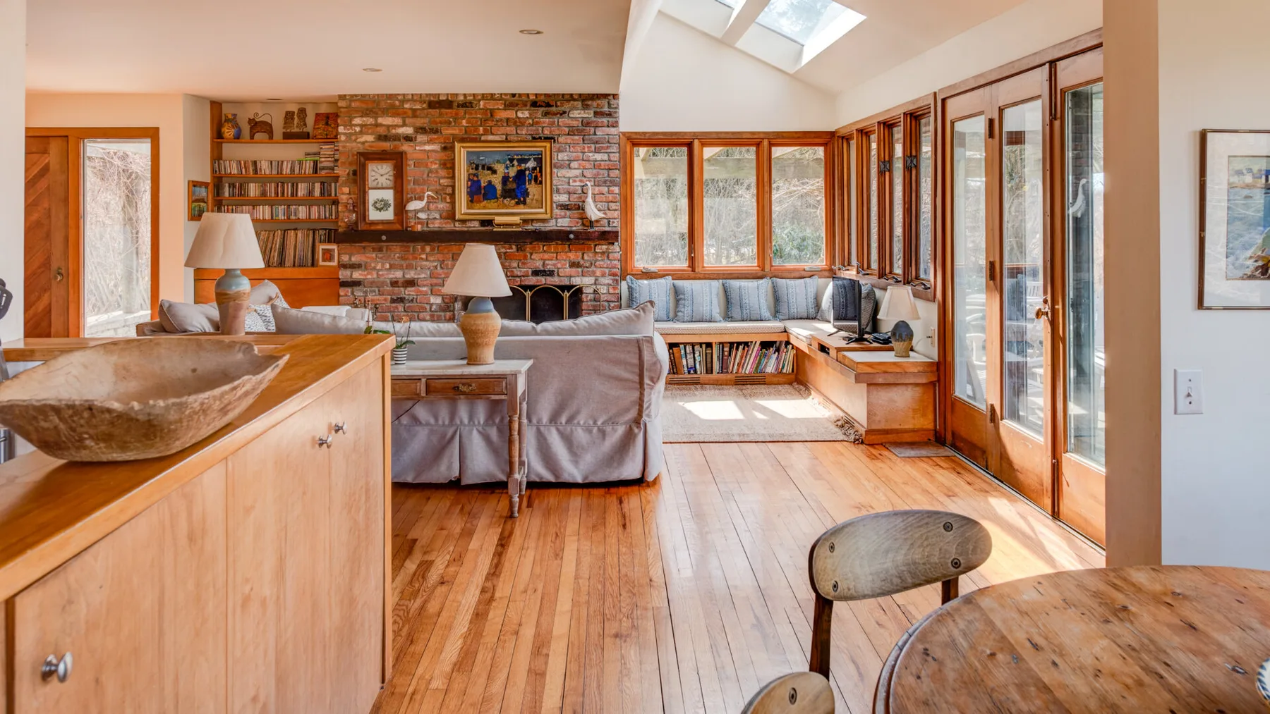 A bright living room with a brick fireplace, large windows, and wood flooring.