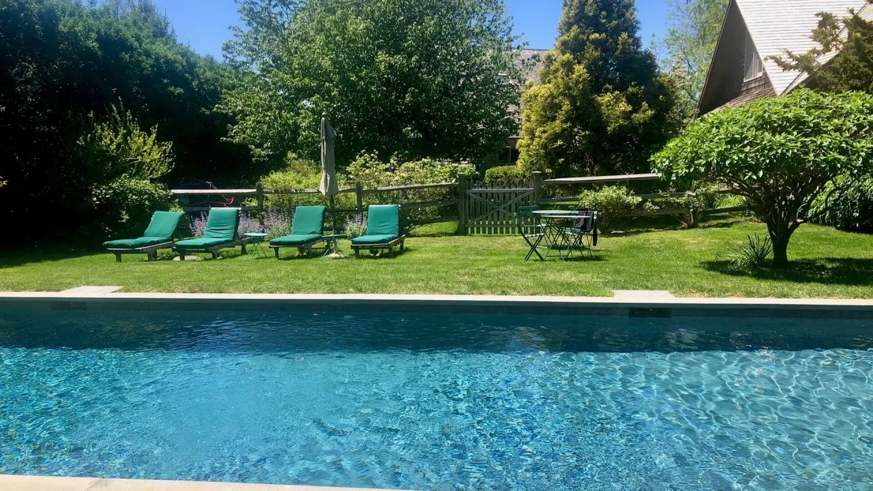 Una piscina al aire libre junto a un césped, asientos de descanso y árboles frondosos.