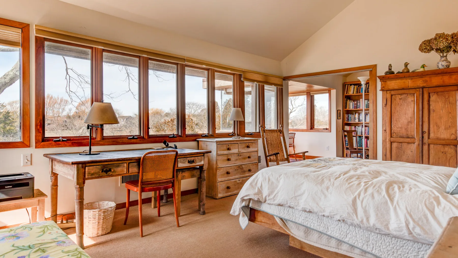 A bedroom with a bed, wooden desk, and large windows overlooking trees.