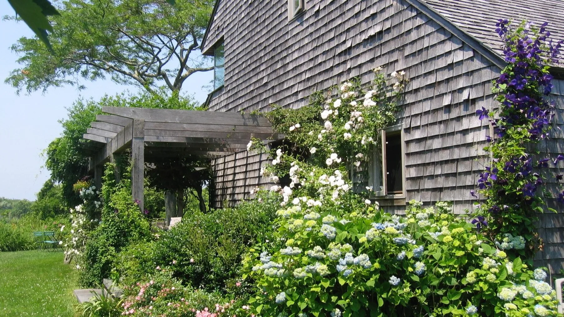 A shingled house beside a wooden pergola, surrounded by flowering garden plants.