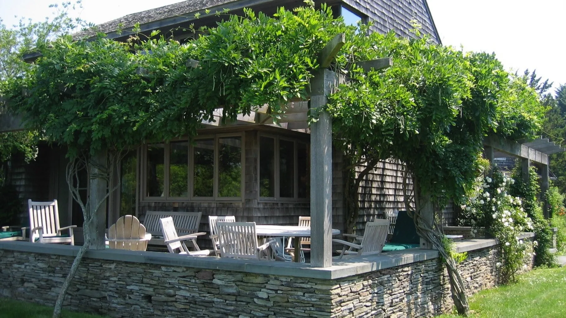 A vine-covered outdoor patio with seating beside a stone retaining wall.