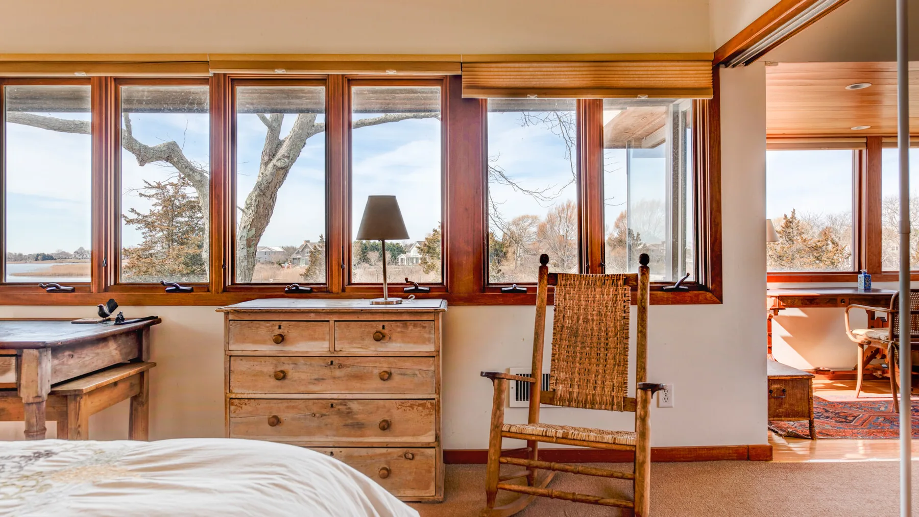 A bedroom with large windows, a wooden dresser, and a rocking chair.
