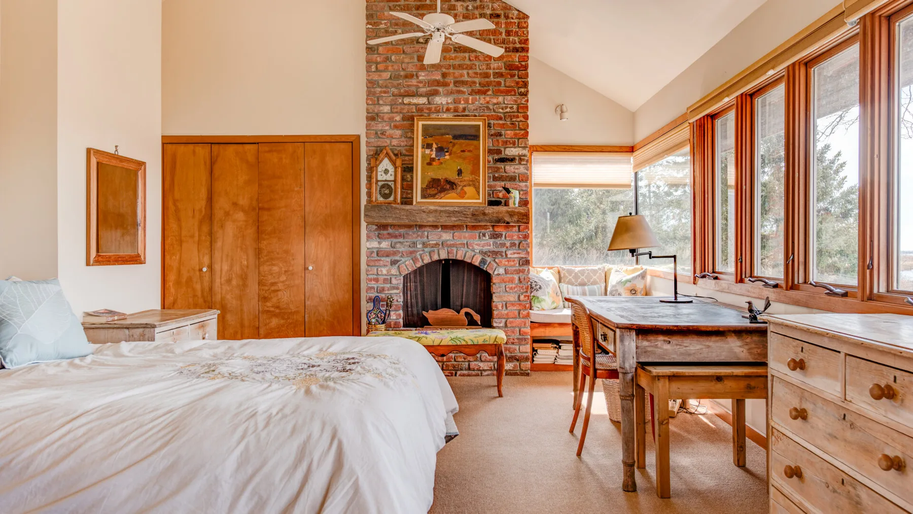 A bright bedroom features a bed, a brick fireplace, and a desk beside large windows.