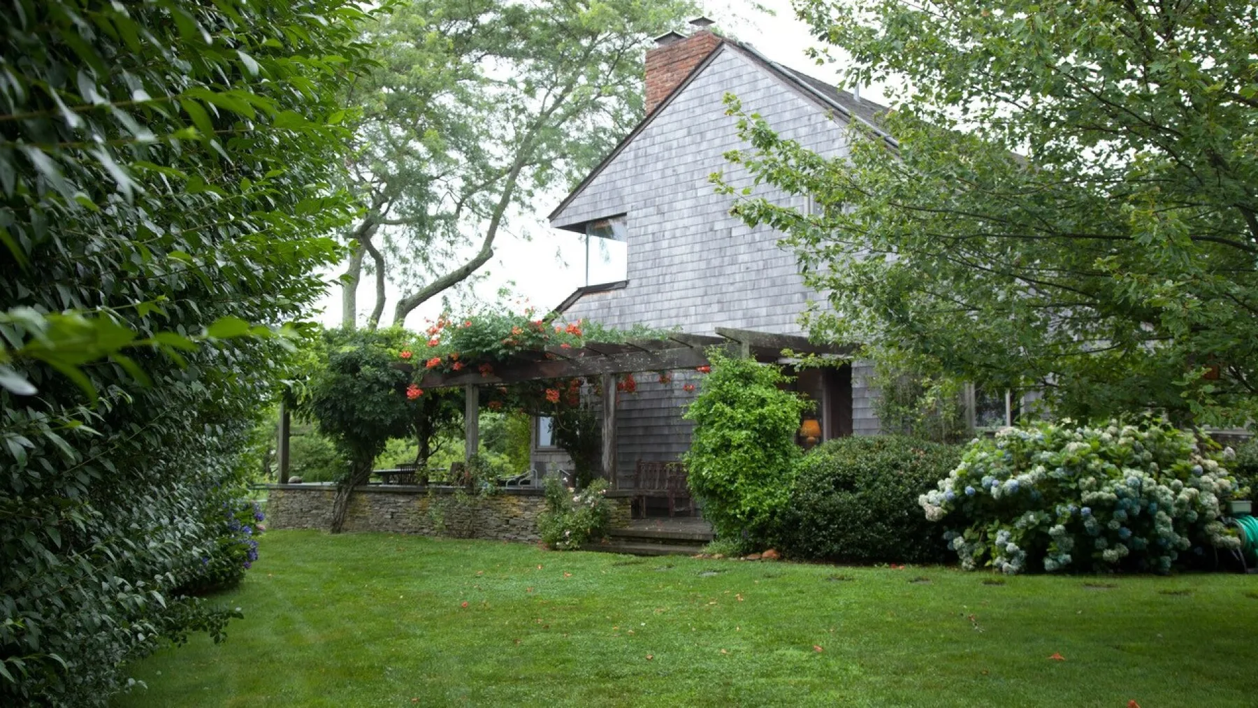 A house stands behind a green lawn, flowering shrubs, and leafy trees.