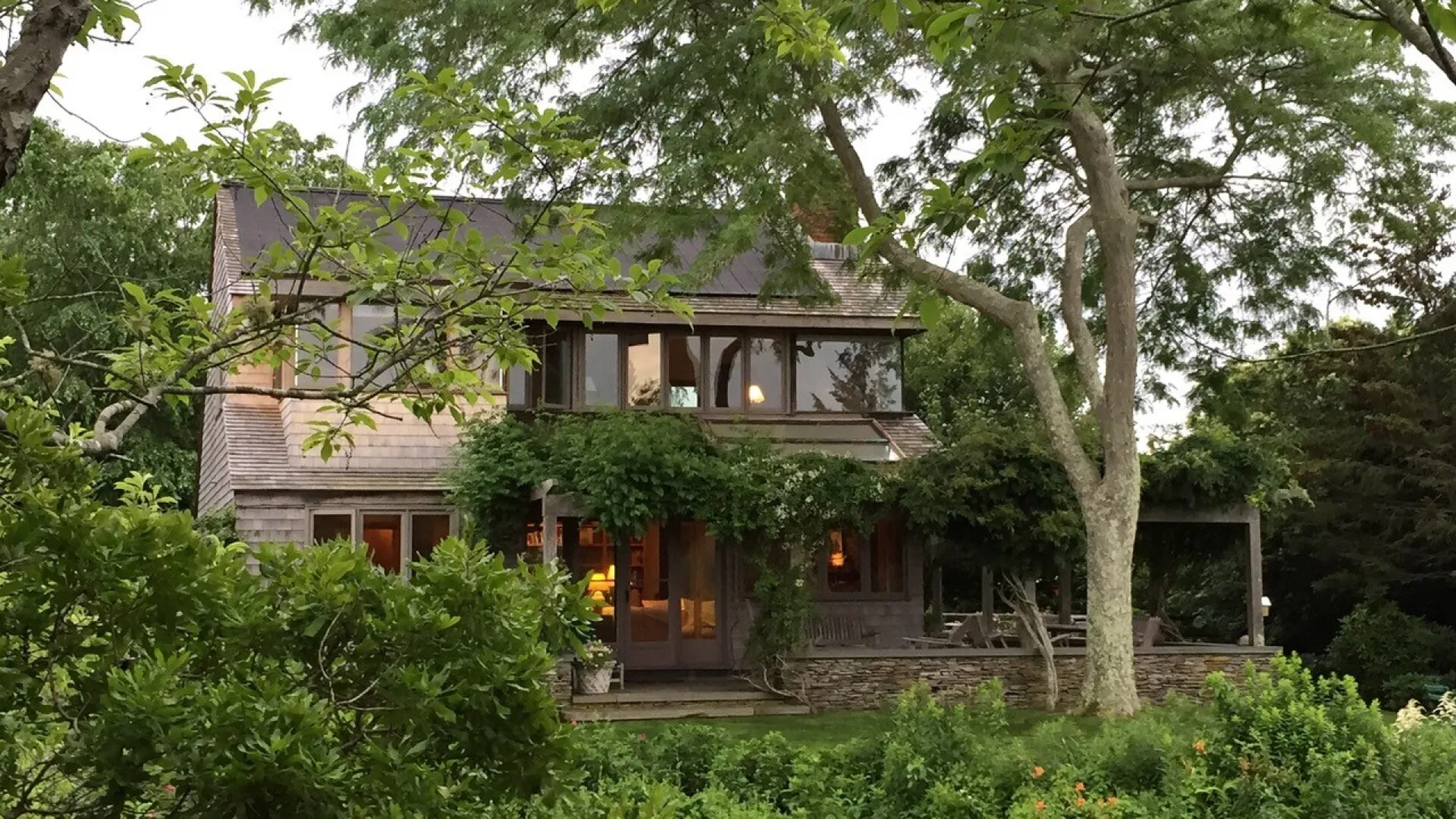 A wooded house exterior is framed by leafy trees and shrubs.