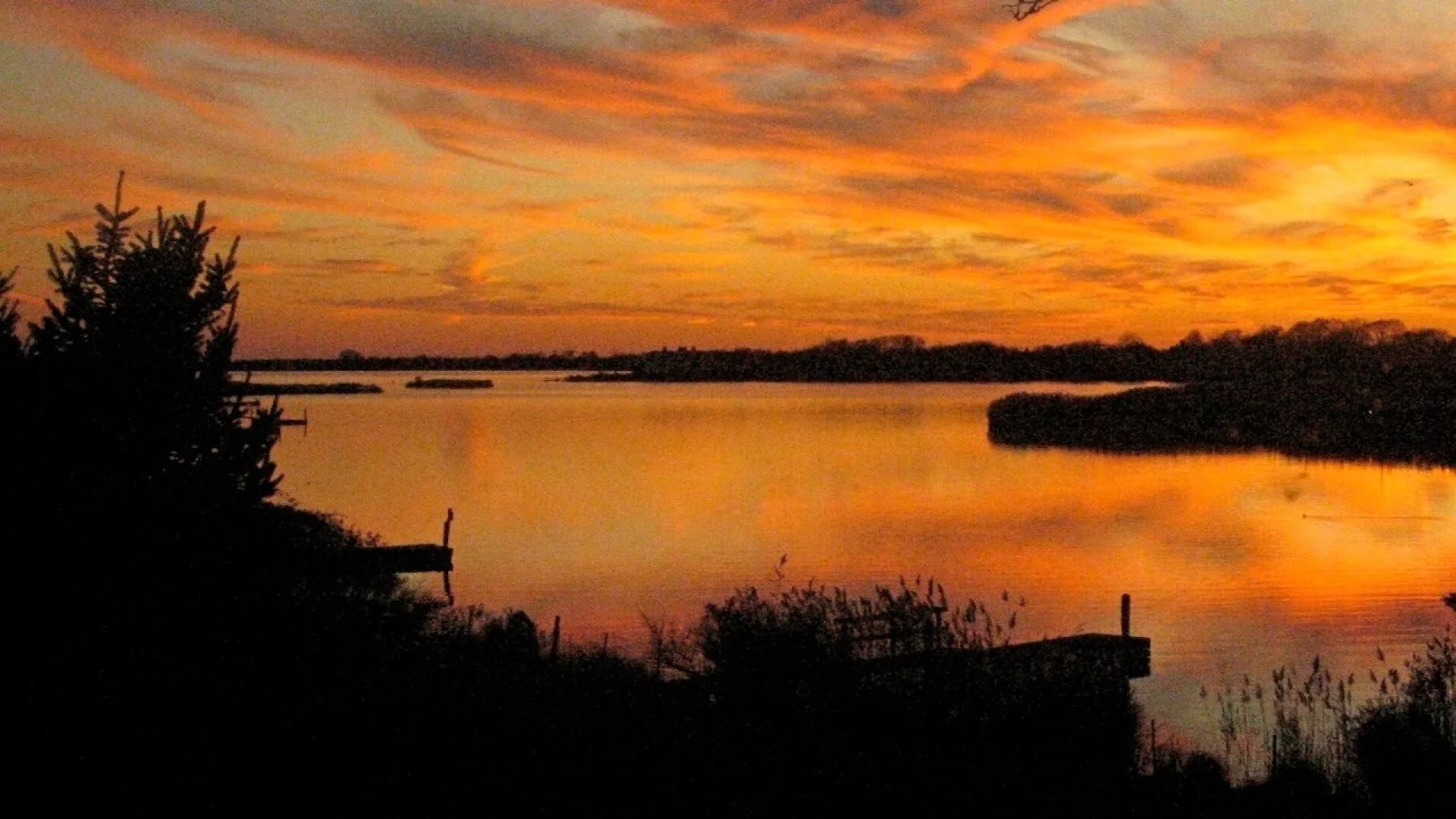 A lake at sunset reflects an orange sky, with dark trees along the shore.