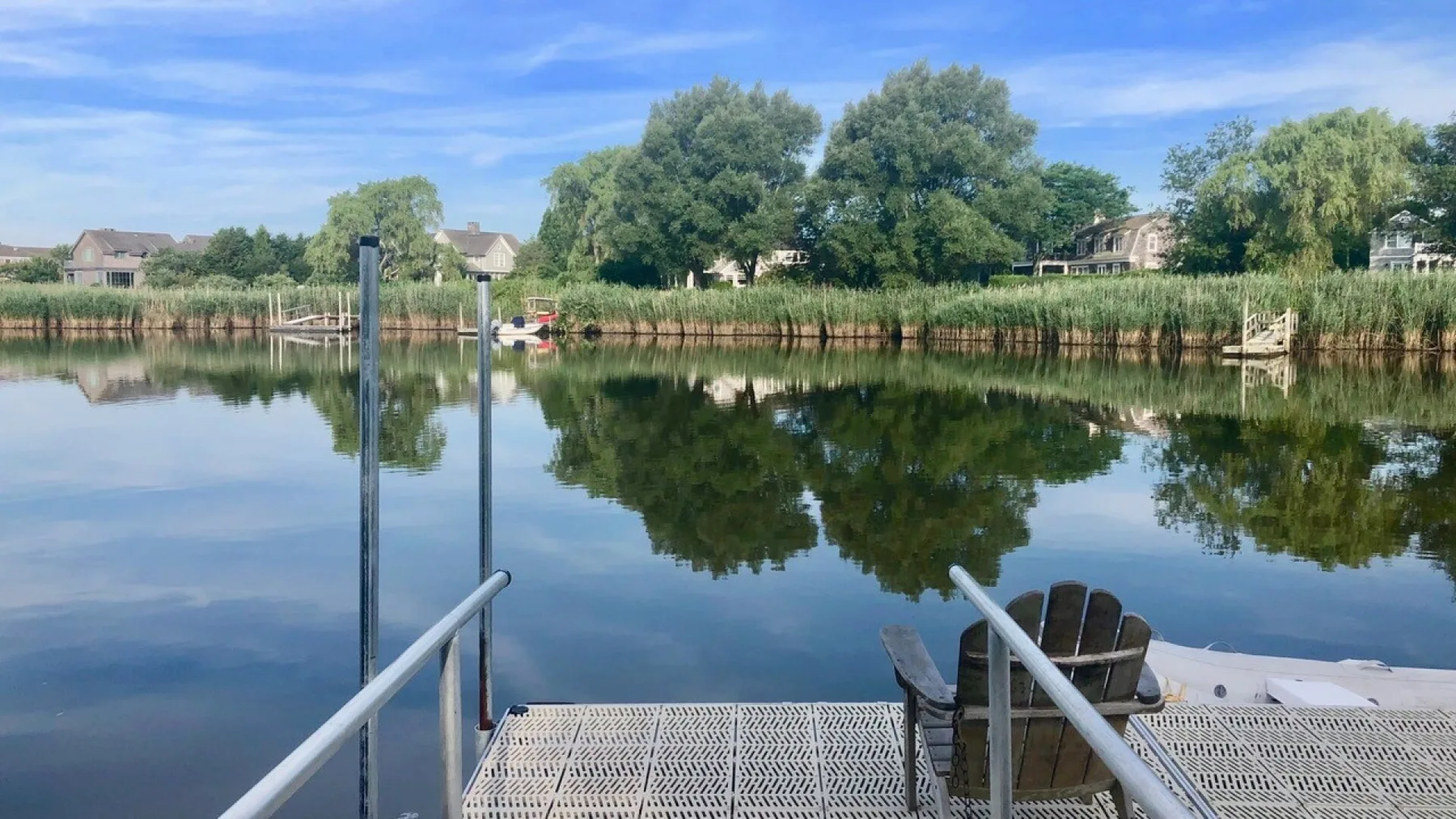 Escena tranquila junto al agua, con un muelle metálico, asientos y árboles reflejados.