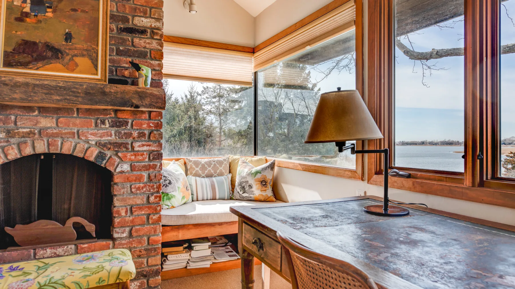 A sunlit sitting area has a brick fireplace, a window seat, and a table.