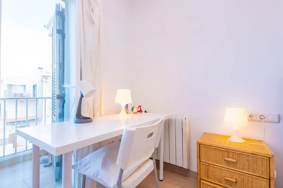 A bright desk area by a large window, with a chair and bedside cabinet.