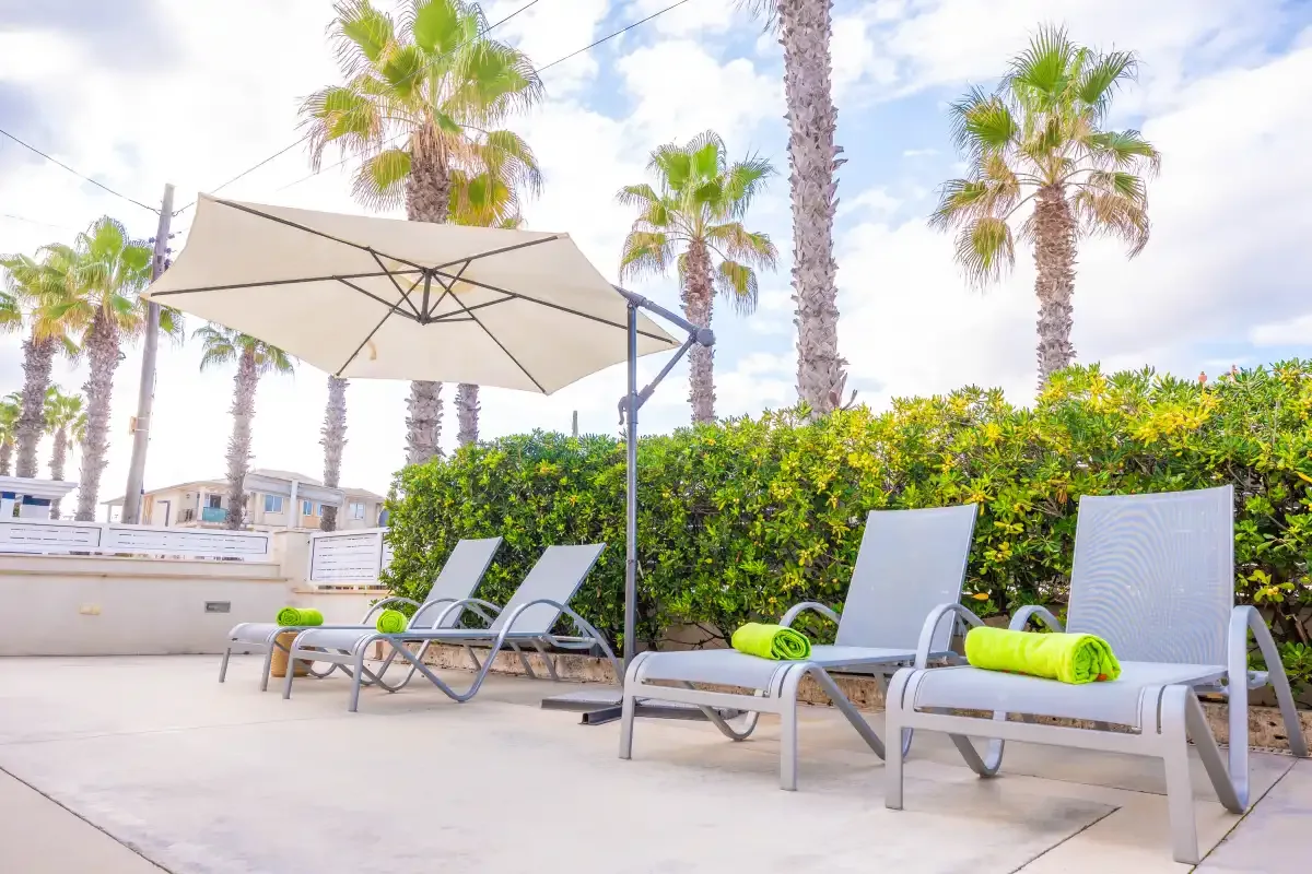 A sunny outdoor poolside patio has lounge seating beneath a large umbrella.