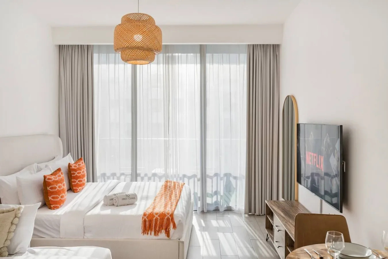 Un dormitorio luminoso con una cama blanca, detalles naranjas y ventanas con cortinas de altura completa.
